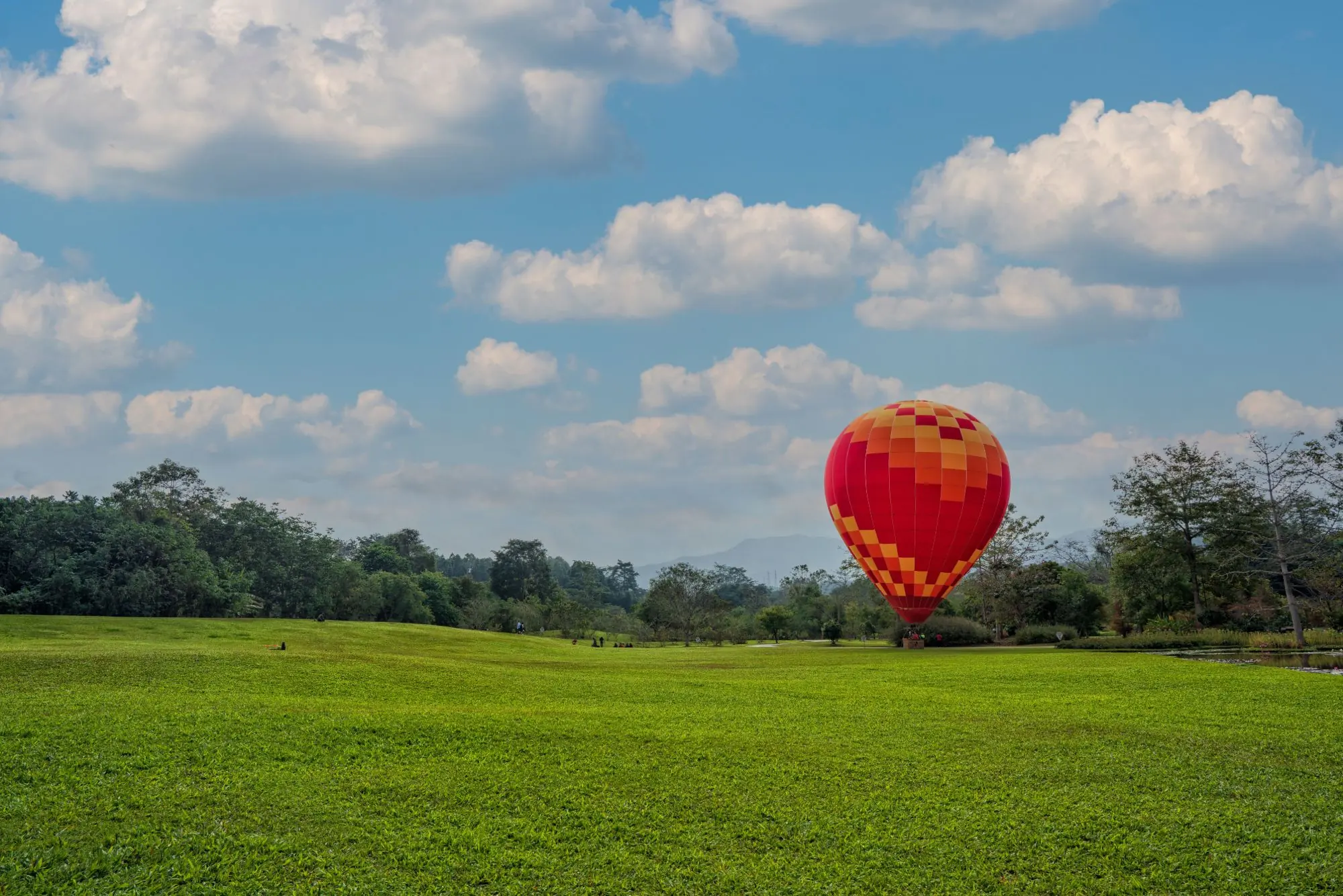 A red and orange hot air balloon standing on a green lawn under a blue sky with white clouds
