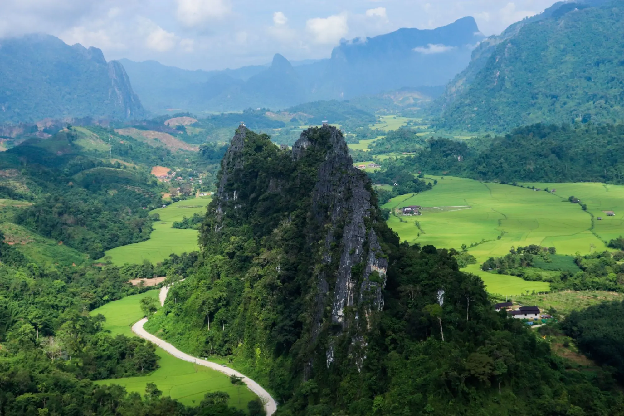 Twin limestone karst peaks rising above bright green rice fields and a winding road in a Laos valley