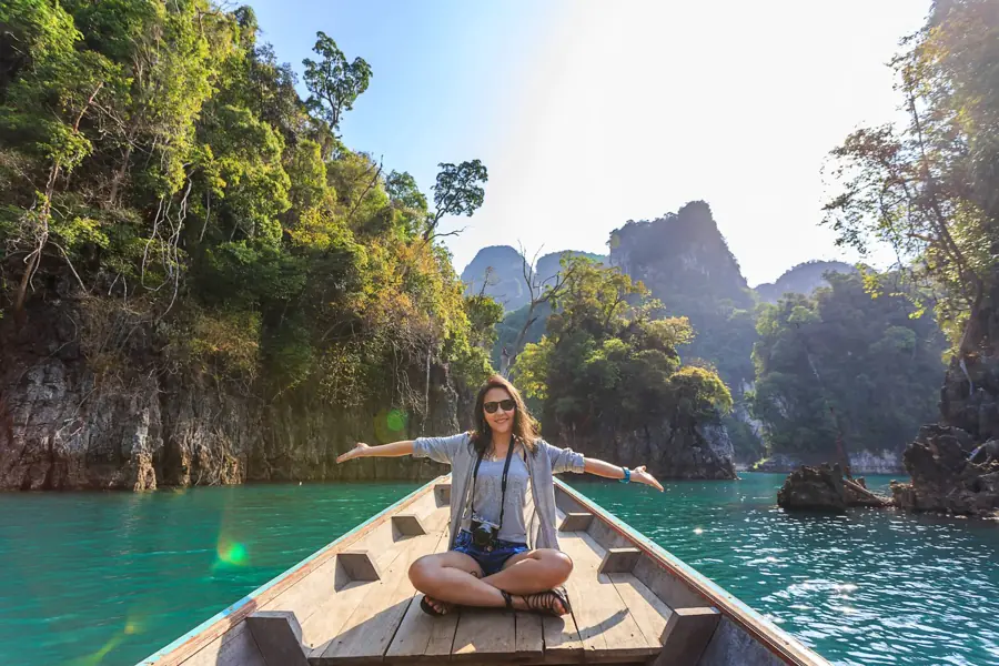 A traveller with open arms on the bow of a longtail boat on a turquoise lake among karst cliffs in Thailand