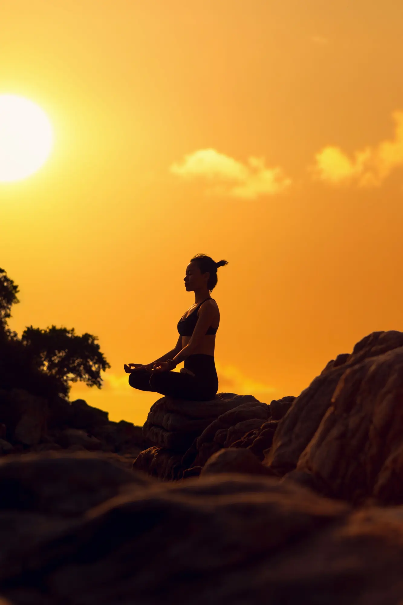 A woman meditating in lotus pose on a rock, silhouetted against a golden sunset
