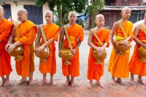 A group of young monks dressed in orange robes standing outdoors, holding alms bowls.